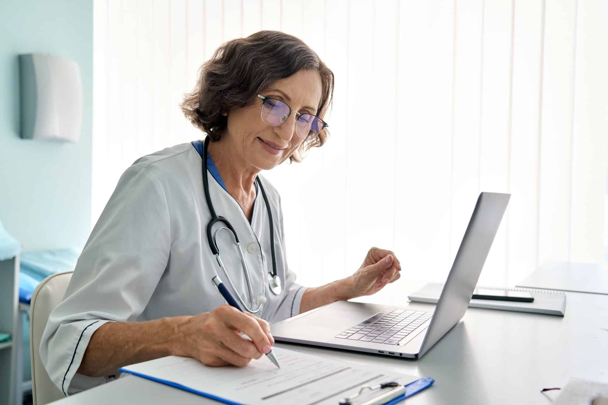 Senior female doctor in clinic consulting remotely using pc laptop computer.
